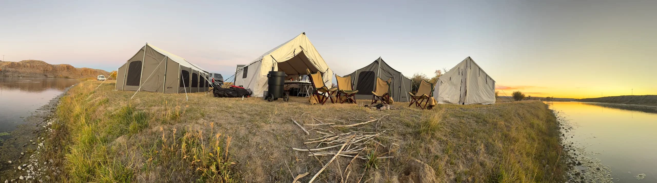 Panorama of a canvas camp on a grassy riverbank at dusk: four canvas tents, camp chairs, and a smoker, with the river on both sides and the sun setting on the right.