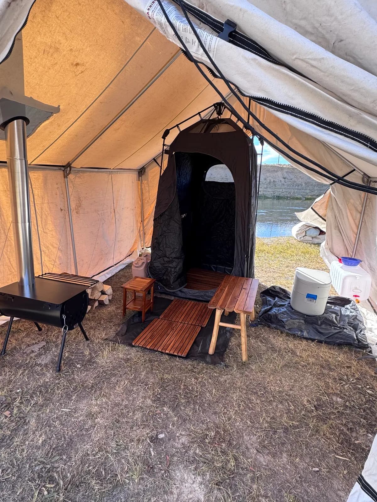 Inside a canvas tent: a shower enclosure on wooden duckboards, a wooden bench, a water jug, and a wood stove.